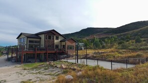 A multi-story house with a balcony, surrounded by a fenced area and a hilly landscape.