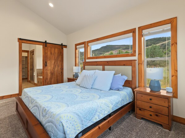 A bedroom with a bed, wooden headboard, bedside tables, and a view of a landscape through windows.
