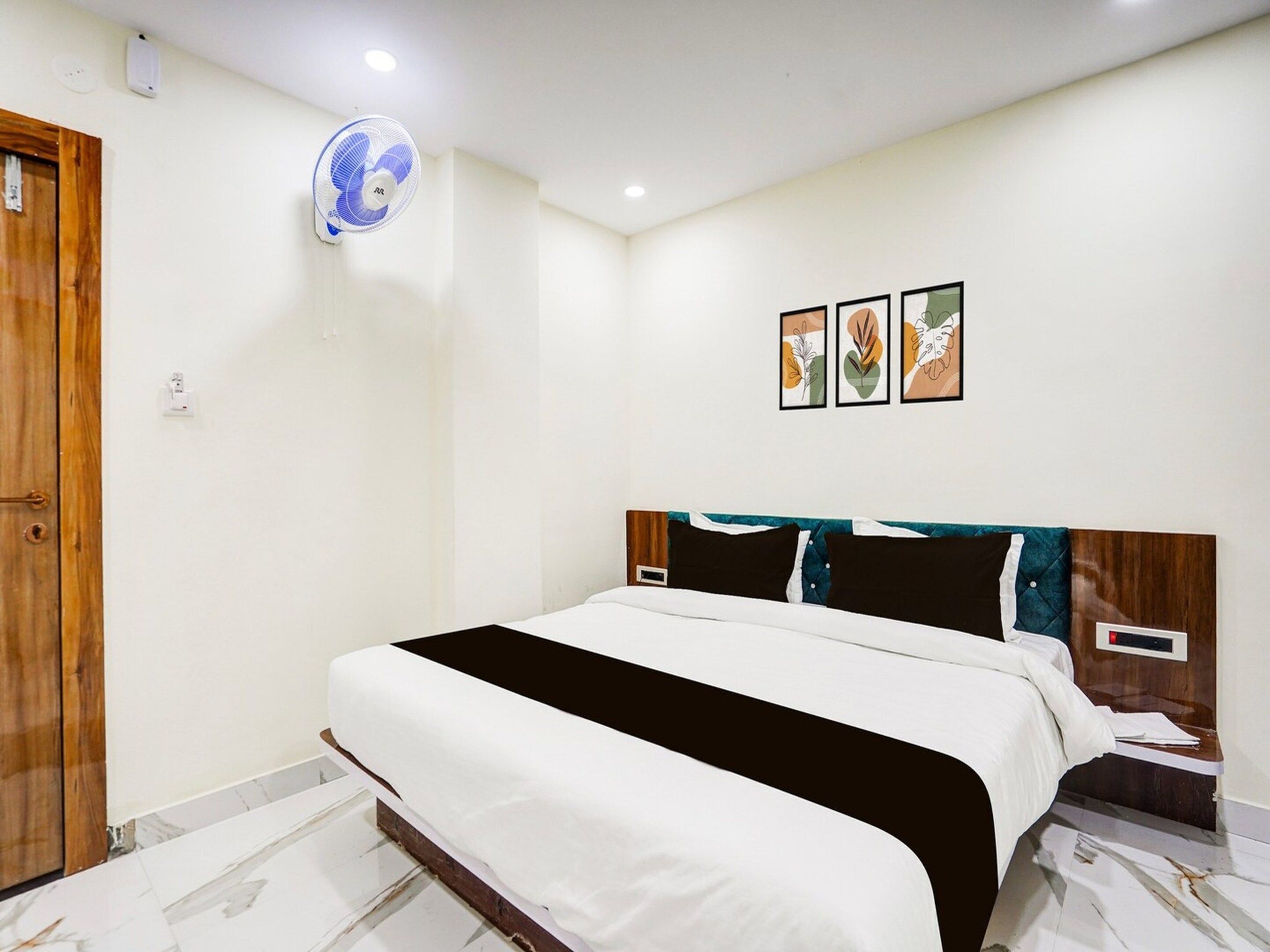A bedroom with a bed, a wooden door, a wall-mounted fan, and three framed pictures.