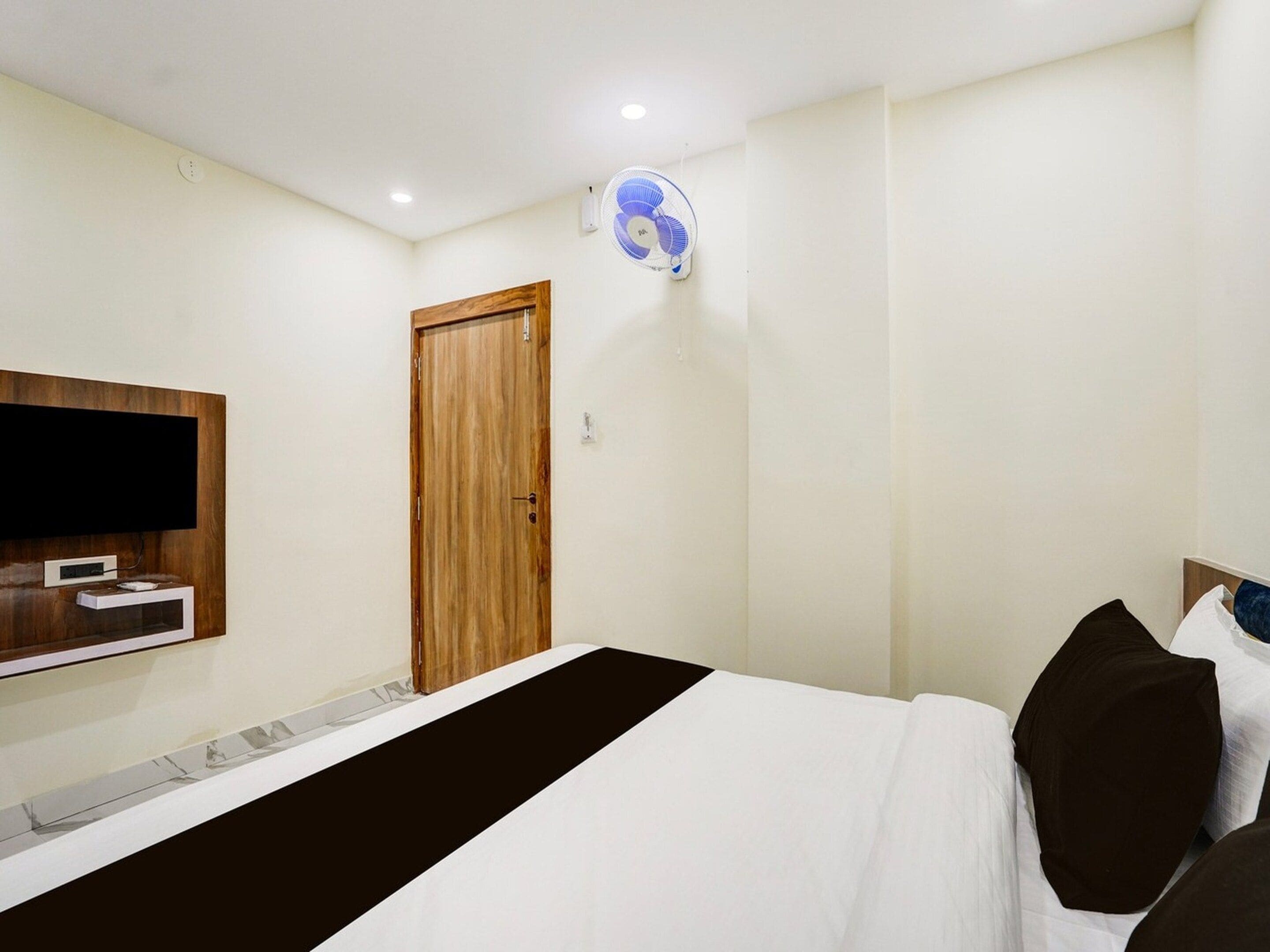 A hotel room with a bed, a TV mounted on the wall, a wooden door, and a ceiling fan.