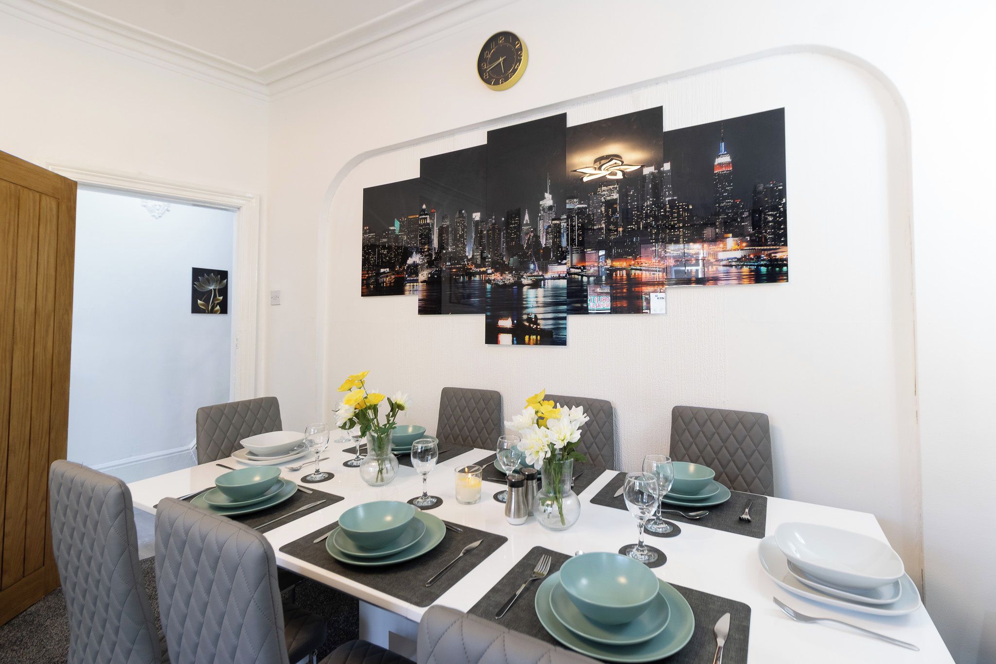 A dining area with a cityscape artwork on the wall, a table set for four with teal plates and glasses, and a vase with yellow flowers.