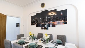 Een eetgedeelte met stadsgezicht kunst aan de muur, een tafel gedekt voor vier met teal borden en glazen, en een vaas met gele bloemen.