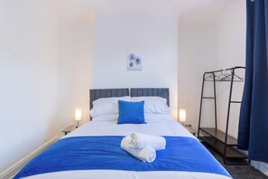 A hotel room with a bed, two bedside lamps, a blue and white bedspread, a rolled white towel, a grey headboard, a wall-mounted artwork, and a black metal drying rack. - Spacious 5 Bedroom House in Manchester City| near Old Trafford| Sleeps up to 13! (Manchester)