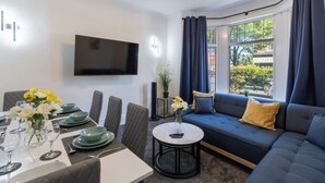 Een moderne woonkamer met een eethoek, een flatscreen televisie aan de muur, een bank met kussens, en een raam met gordijnen.