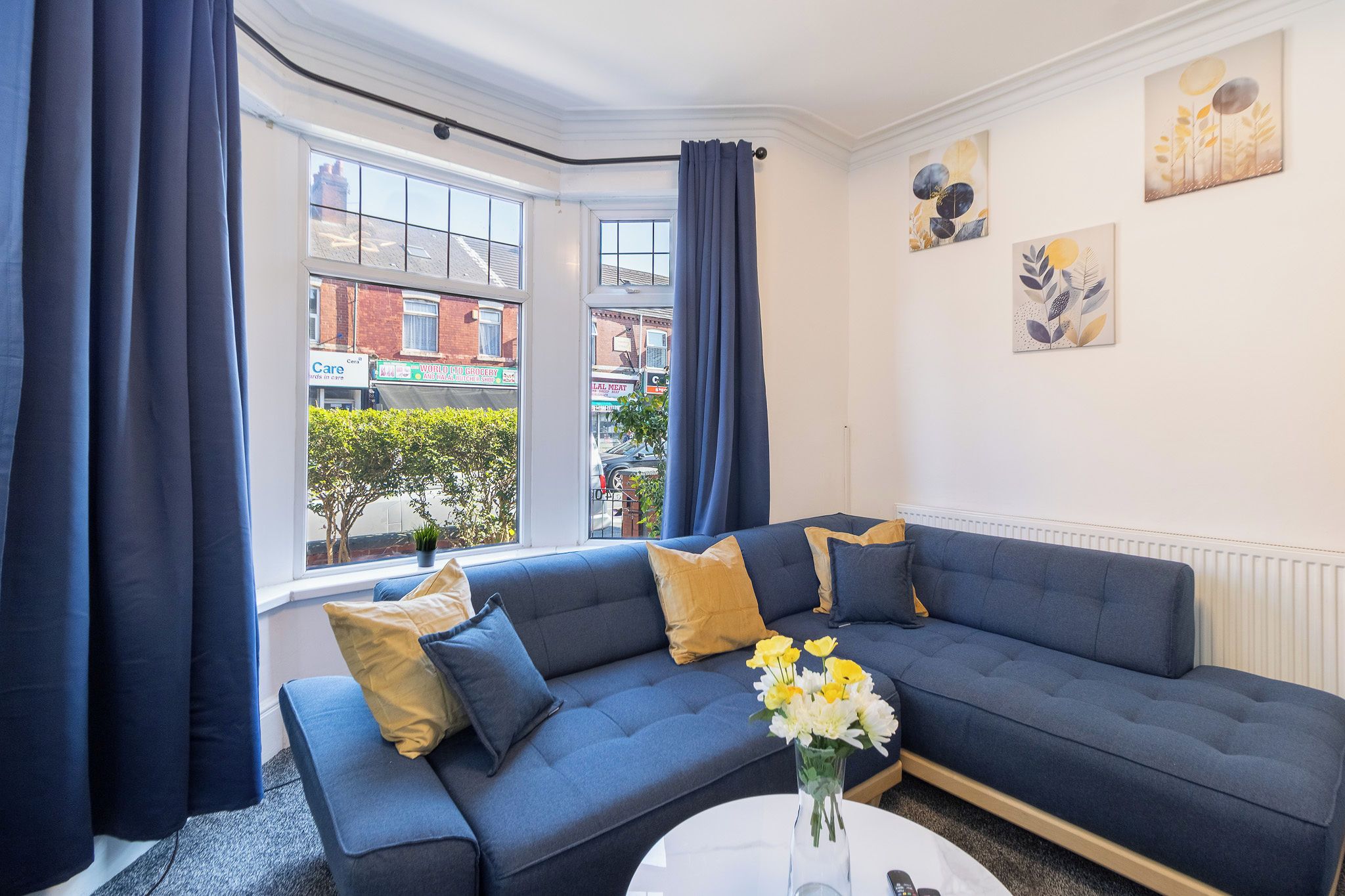 A modern living room with a blue sofa, a white coffee table, and a view of the street through large windows.