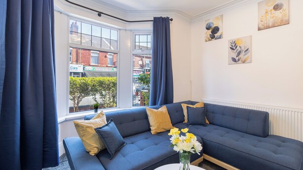 Een moderne woonkamer met een blauwe bank, een witte salontafel en uitzicht op de straat door grote ramen.