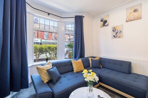 A modern living room with a blue sofa, a white coffee table, and a view of the street through large windows. - Spacious 5 Bedroom House in Manchester City| near Old Trafford| Sleeps up to 13! (Manchester)