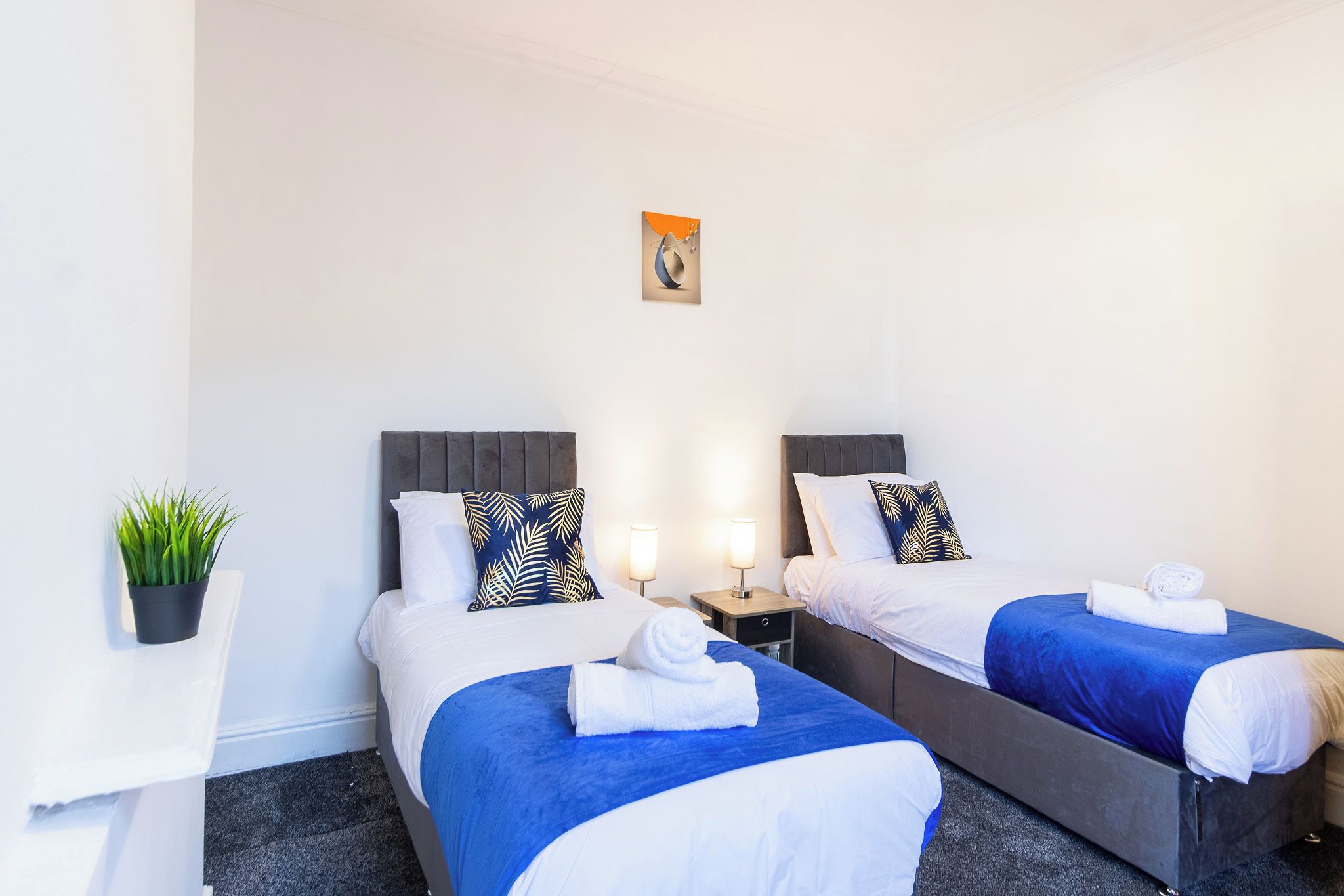 A hotel room with two beds, each with a blue bedspread and white pillows, a small table with a plant, and a wall-mounted artwork.