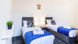 Een hotelkamer met twee bedden, elk met een blauwe spreidek en witte kussens, een kleine tafel met een plant erop, en een kunstwerk aan de muur.