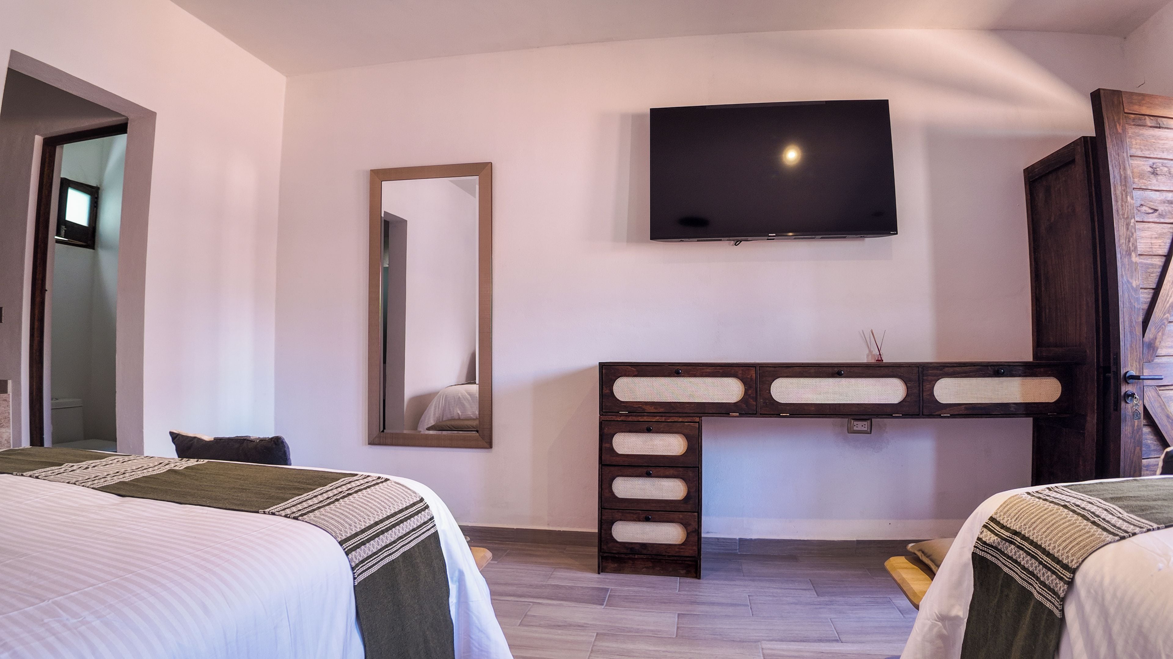 Habitación de hotel con dos camas, televisor montado en la pared, un espejo y un aparador de madera con cajones.