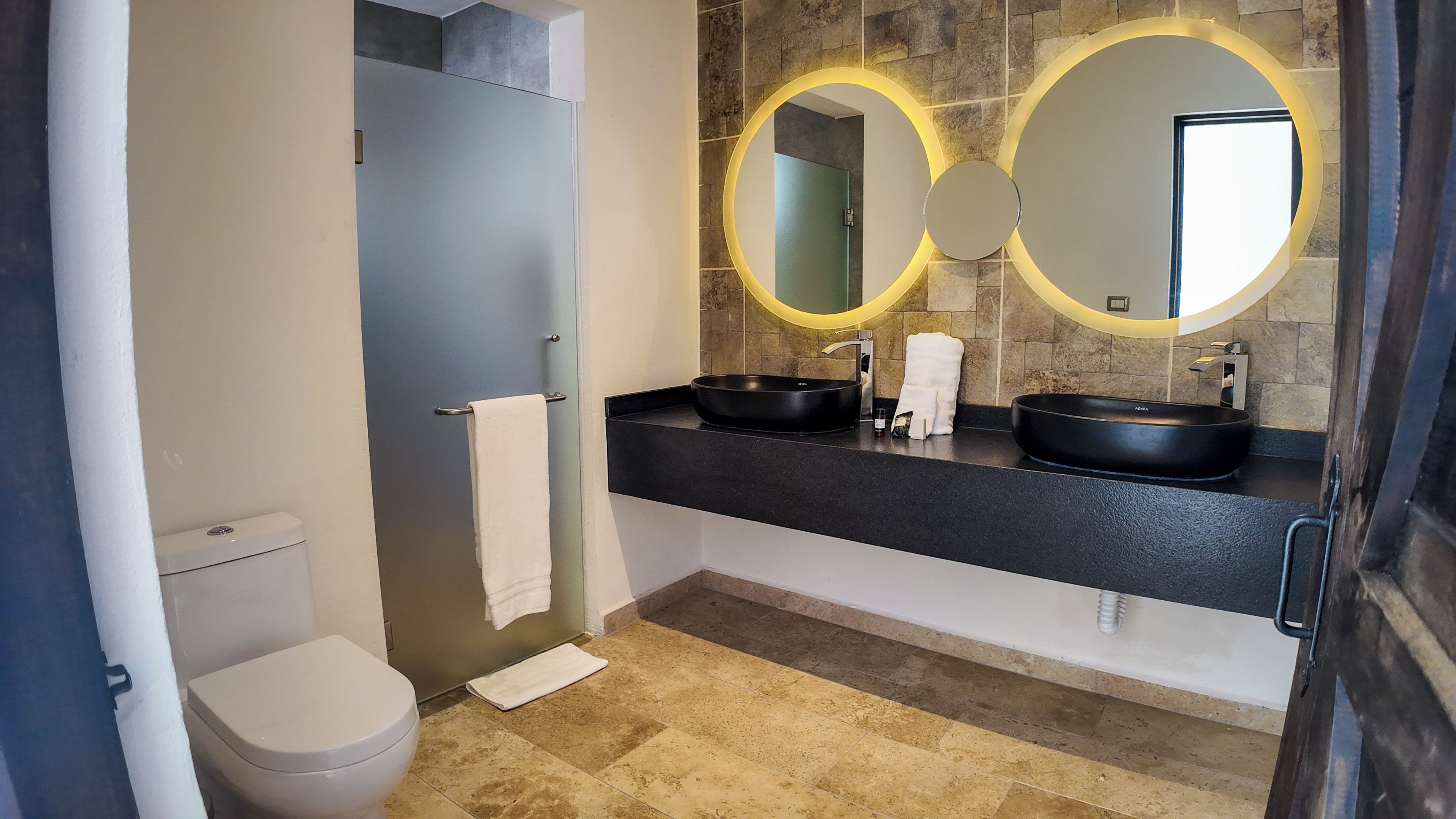A modern bathroom with a toilet, two sinks, and a large mirror with circular lighting.