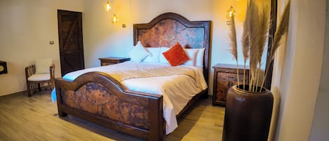 Quarto com cama de madeira, mesa de cabeceira, cadeira e um vaso grande com capim-do-campo.
