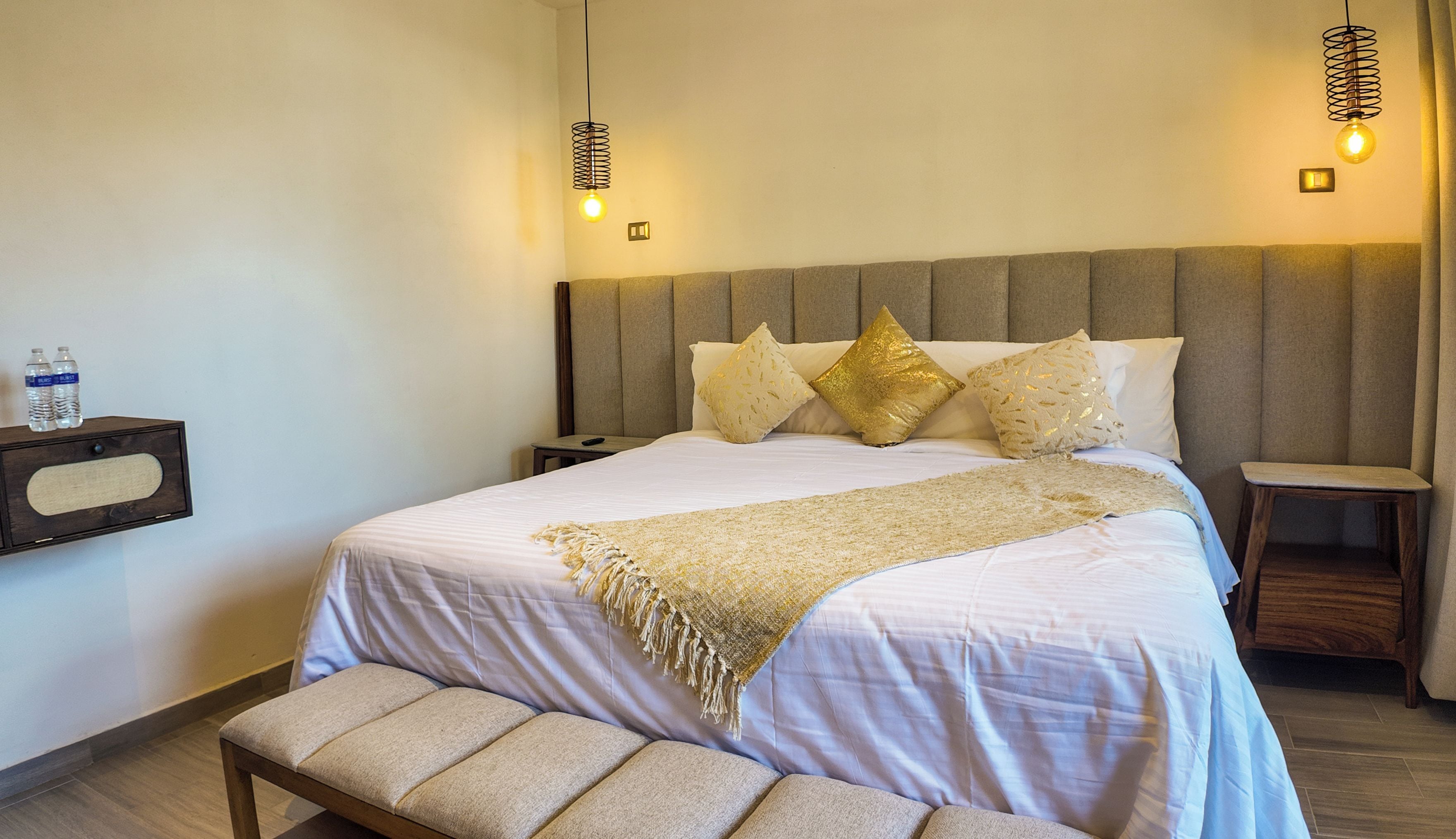 A neatly made bed with a headboard, a bedside table, a bench, and two wall-mounted lamps.