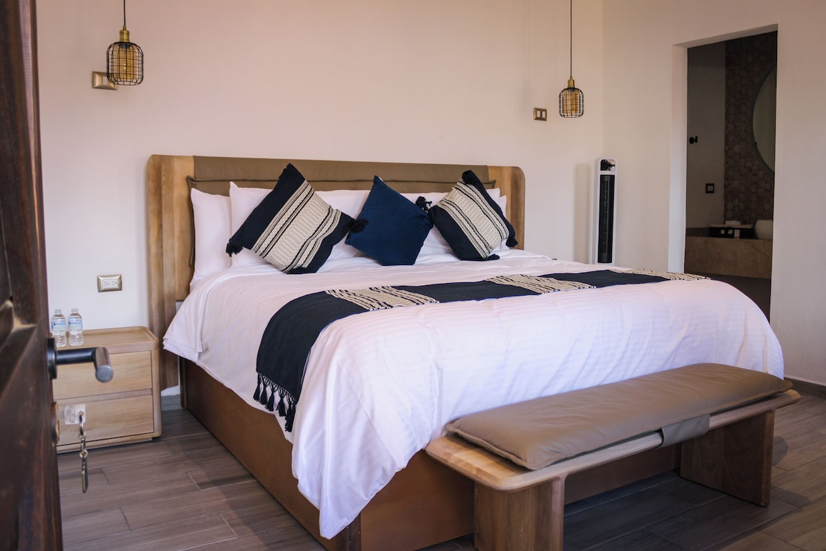 Ein ordentlich bezogenes Bett mit Holz-Kopfteil, ein Nachttisch mit Lampe und eine Bank am Fußende.