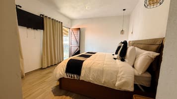 A bedroom with a bed, curtains, a wooden floor, and a view through a door.