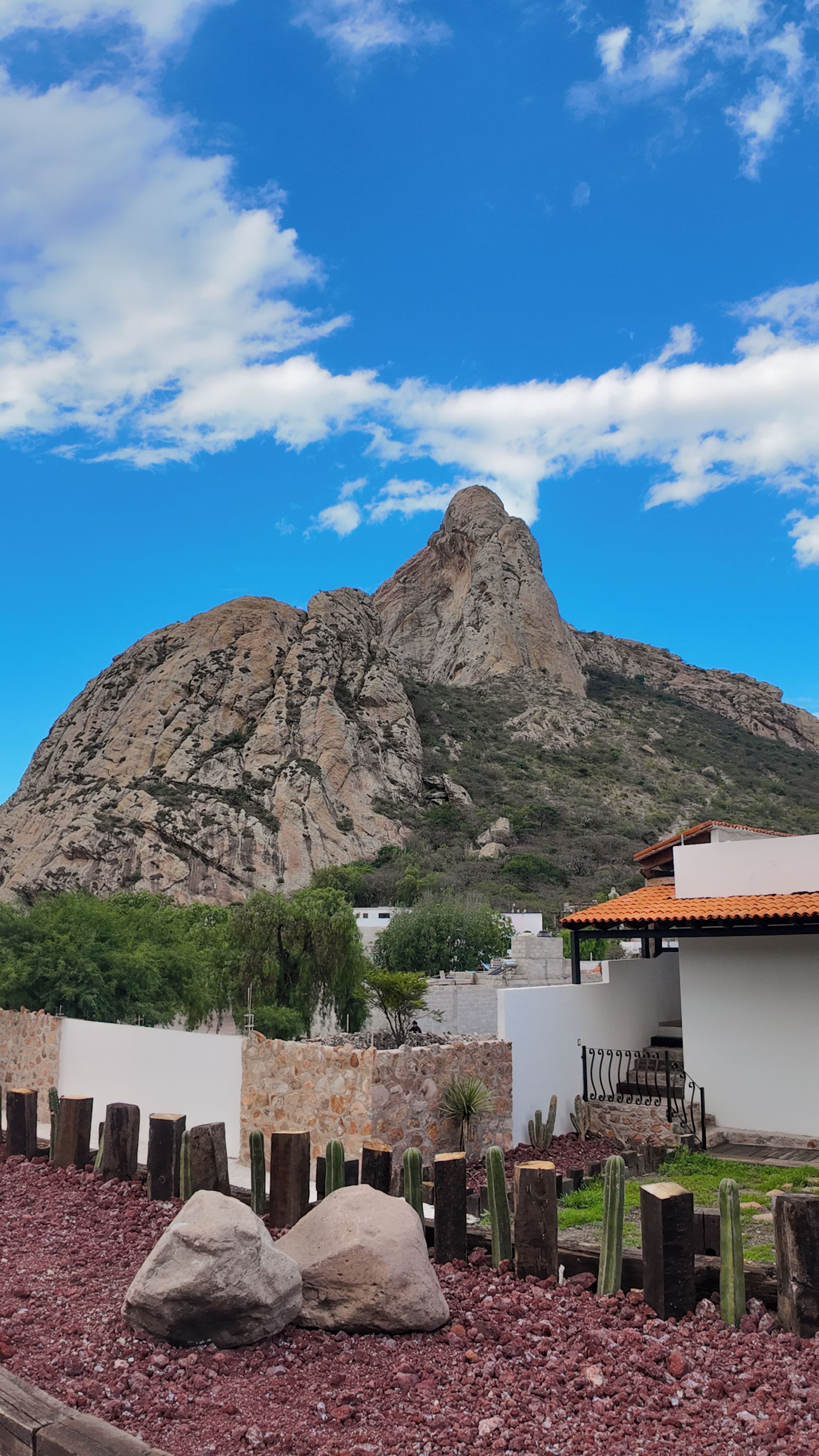 A mountain with a rocky peak, a white building with a red roof, and a garden with cacti and rocks.