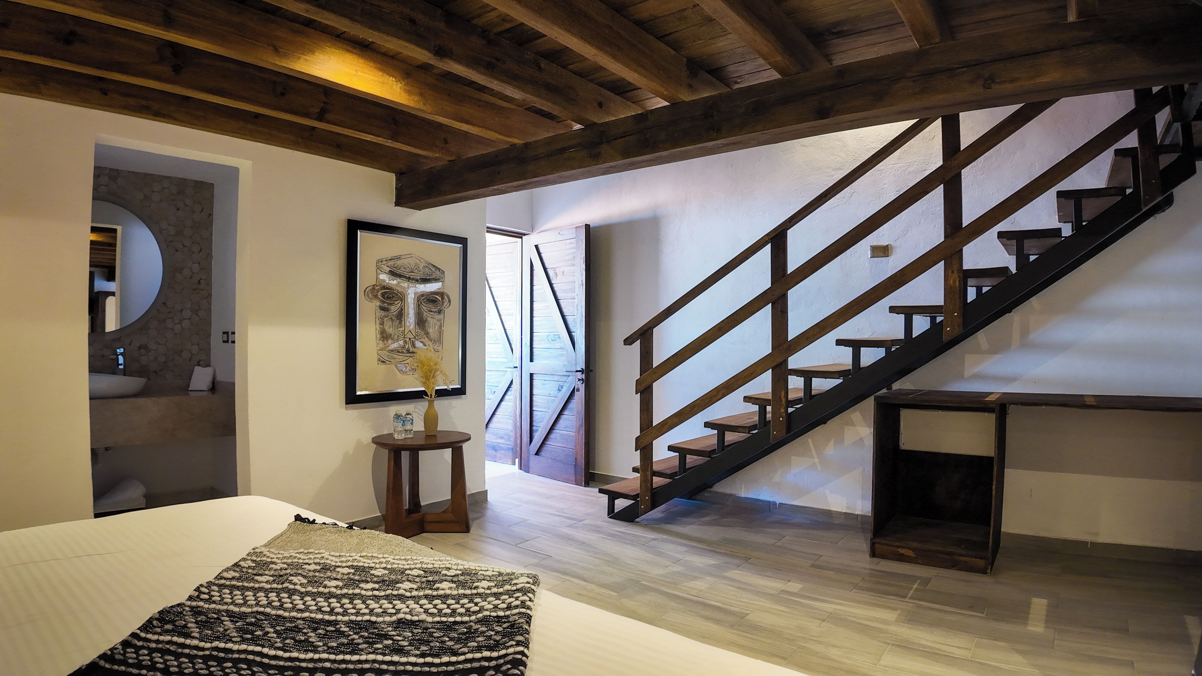 A bedroom with a bed, a wooden staircase, a round mirror, and a painting on the wall.