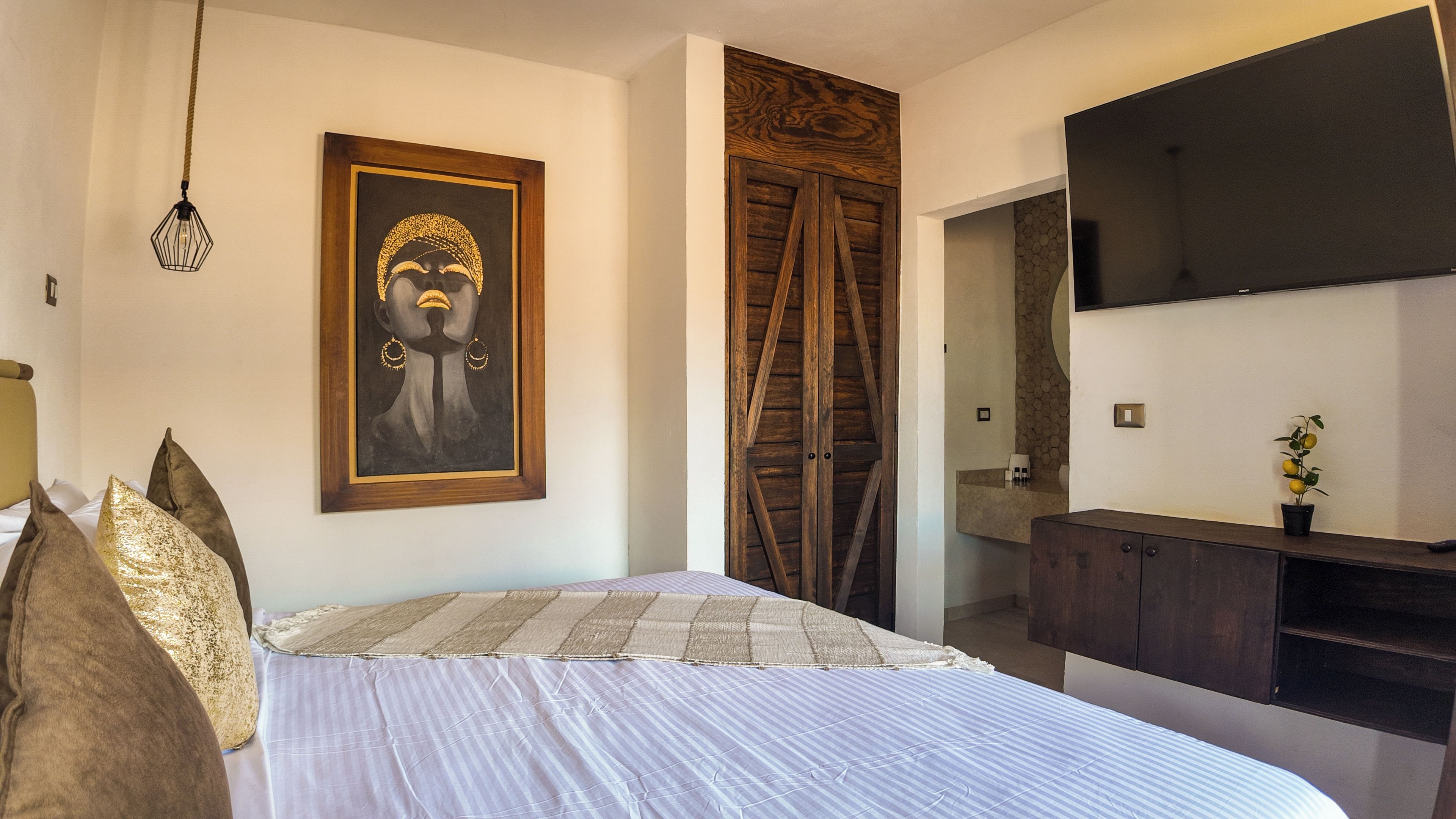 Un dormitorio con cama, una obra de arte enmarco, un armario de madera, televisor y una planta en maceta.