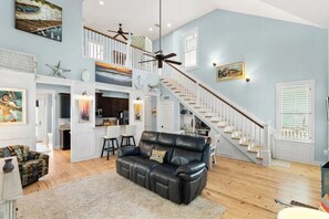 A spacious living area with a black leather sofa, wooden flooring, a staircase with white railings, and a kitchen area in the background. - Where the Boardwalk meets the Bay! (Rockport)