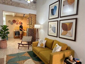 A living room with a sofa, a chair, a potted plant, and a dining area with a person cooking. - Authentic Downtown Charm with Plunge Pool & Fast Wi-Fi | Long-Stay Discount (Mérida)