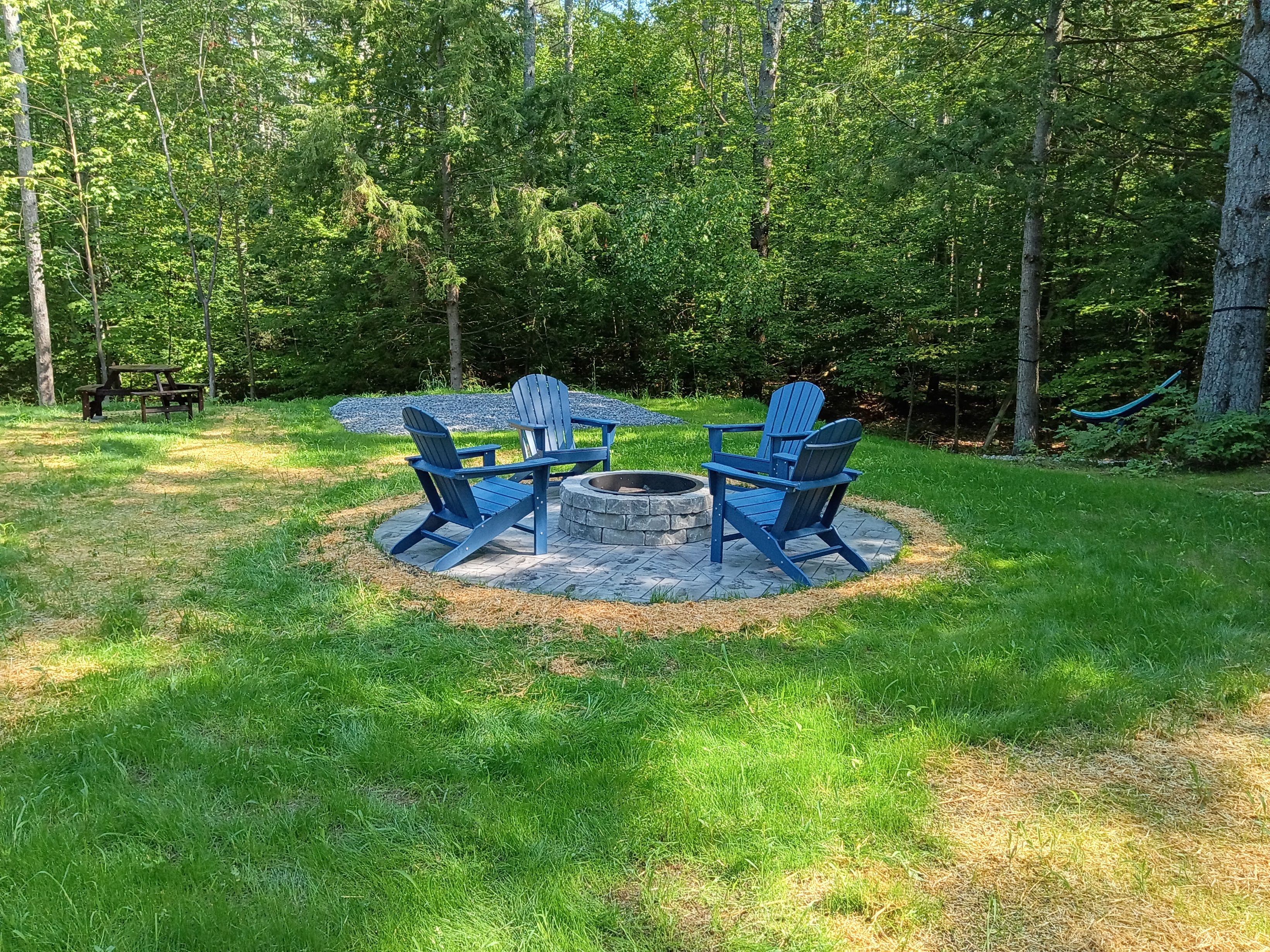 Un espace extérieur circulaire aménagé avec des sièges, doté d’un foyer central entouré d’un anneau de pierres, et comprenant deux fauteuils Adirondack.