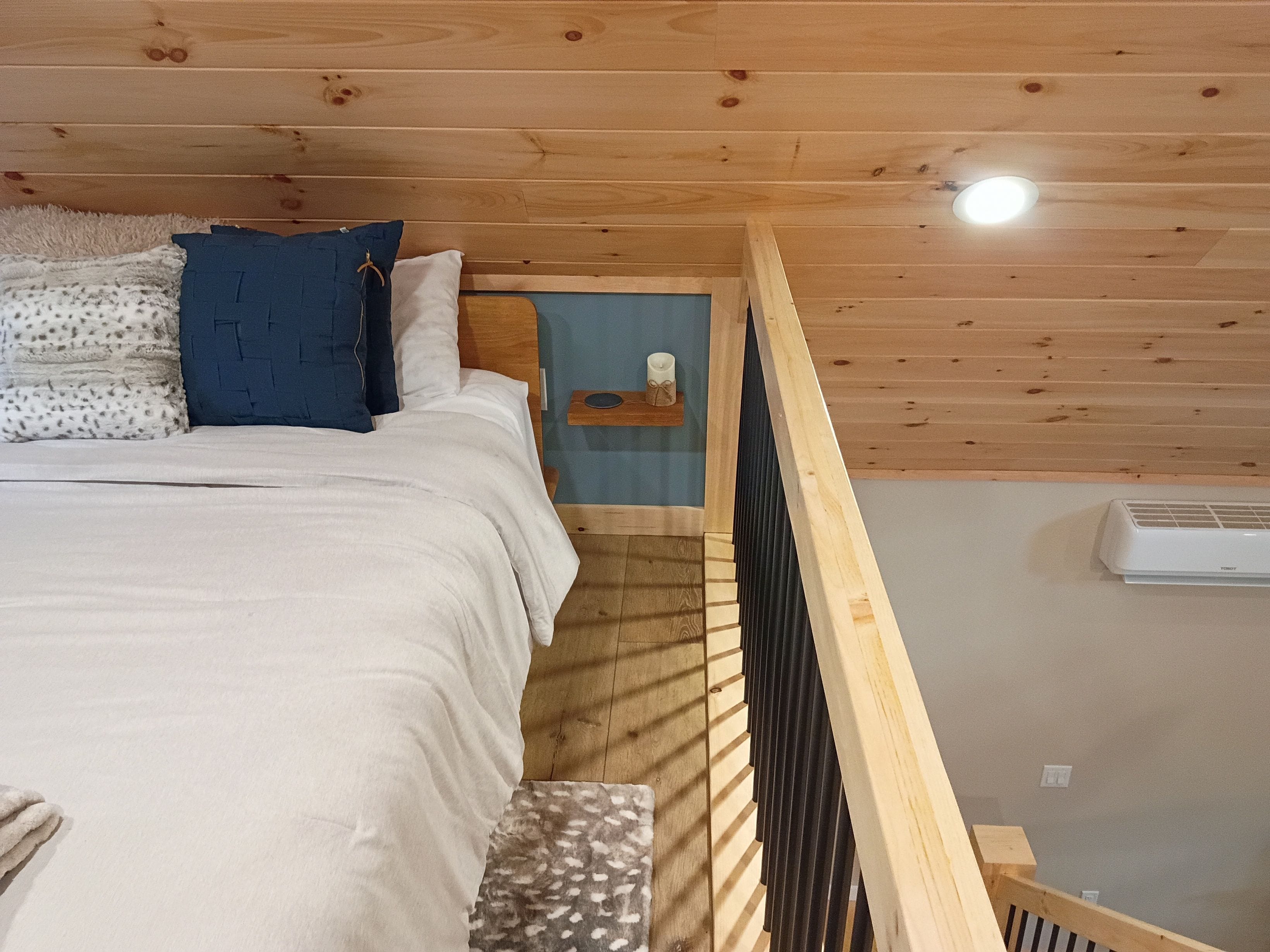 Een slaapkamer met een houten plafond, een bed met wit en gestreept beddengoed, een blauw kussen en een klein nachtkastje met een kaars.