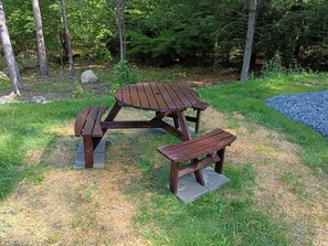 Ein Picknicktisch mit Bänken in einer grünen Umgebung, im Hintergrund Bäume.