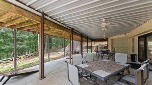 Une terrasse extérieure couverte, dotée d’une table et de chaises, d’un ventilateur de plafond et offrant une vue sur la forêt environnante.