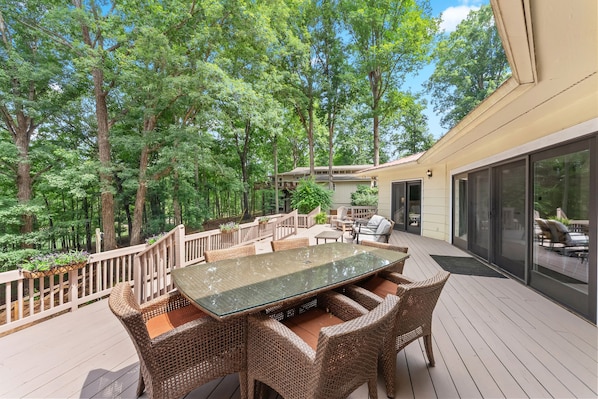 Une terrasse extérieure dotée d’une table à plateau de verre et de chaises en osier, entourée d’une balustrade en bois et d’une végétation luxuriante.