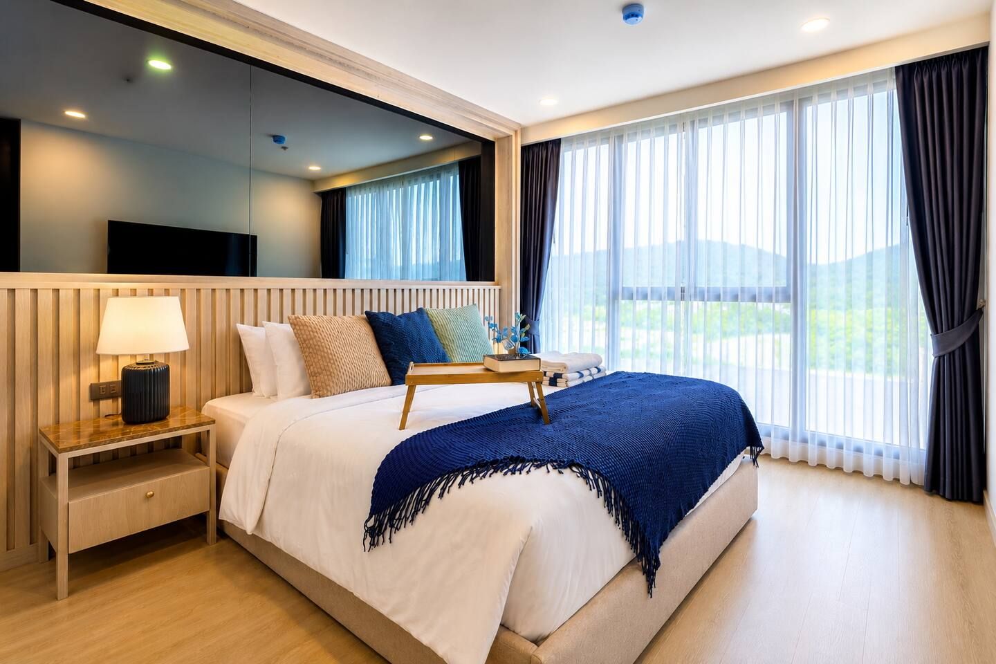 A hotel room with a bed, bedside table, lamp, and a view of the outdoors.