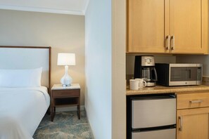 Habitació d'hotel amb un llit, una tauleta de nit amb una làmpada, una cafetera, un microones i un frigorífic.