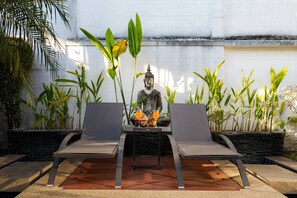 Eine Buddha-Statue sitzt zwischen zwei Liegestühlen, vor ihr steht ein Tisch, umgeben von tropischen Pflanzen.