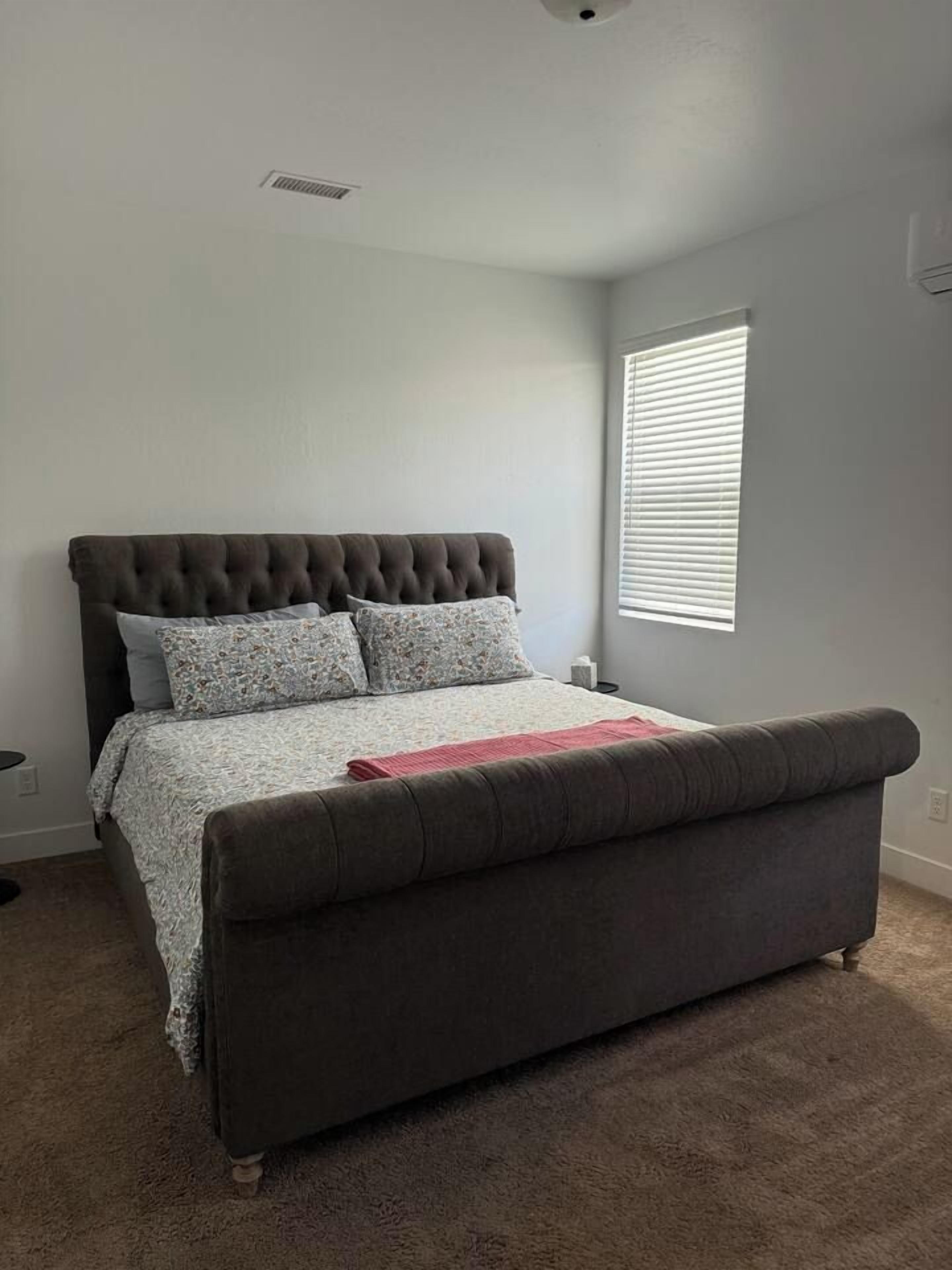 Quarto com cama, janela com persianas, ventilação no teto e piso coberto por carpete.