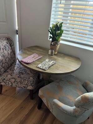 Un comedor con una mesa redonda de madera, dos sillas estampadas, una pequeña planta en maceta y una ventana con persianas.