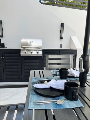 Een buitenkeuken met een barbecue, een tafel gedekt met een bord, een lepel, een vork en een mok.