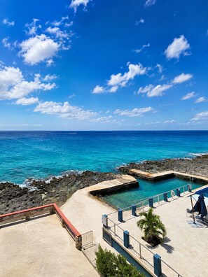 A coastal view with a clear sky, a rocky shoreline, and a swimming pool area. - Amazing Oceanfront Condo w Onsite Diving and Restaurant (West Bay)
