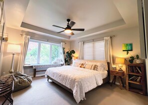 Une chambre à coucher comprenant un lit, un ventilateur de plafond, une fenêtre avec des rideaux, une lampe de chevet, une table de chevet, une bibliothèque et une plante.