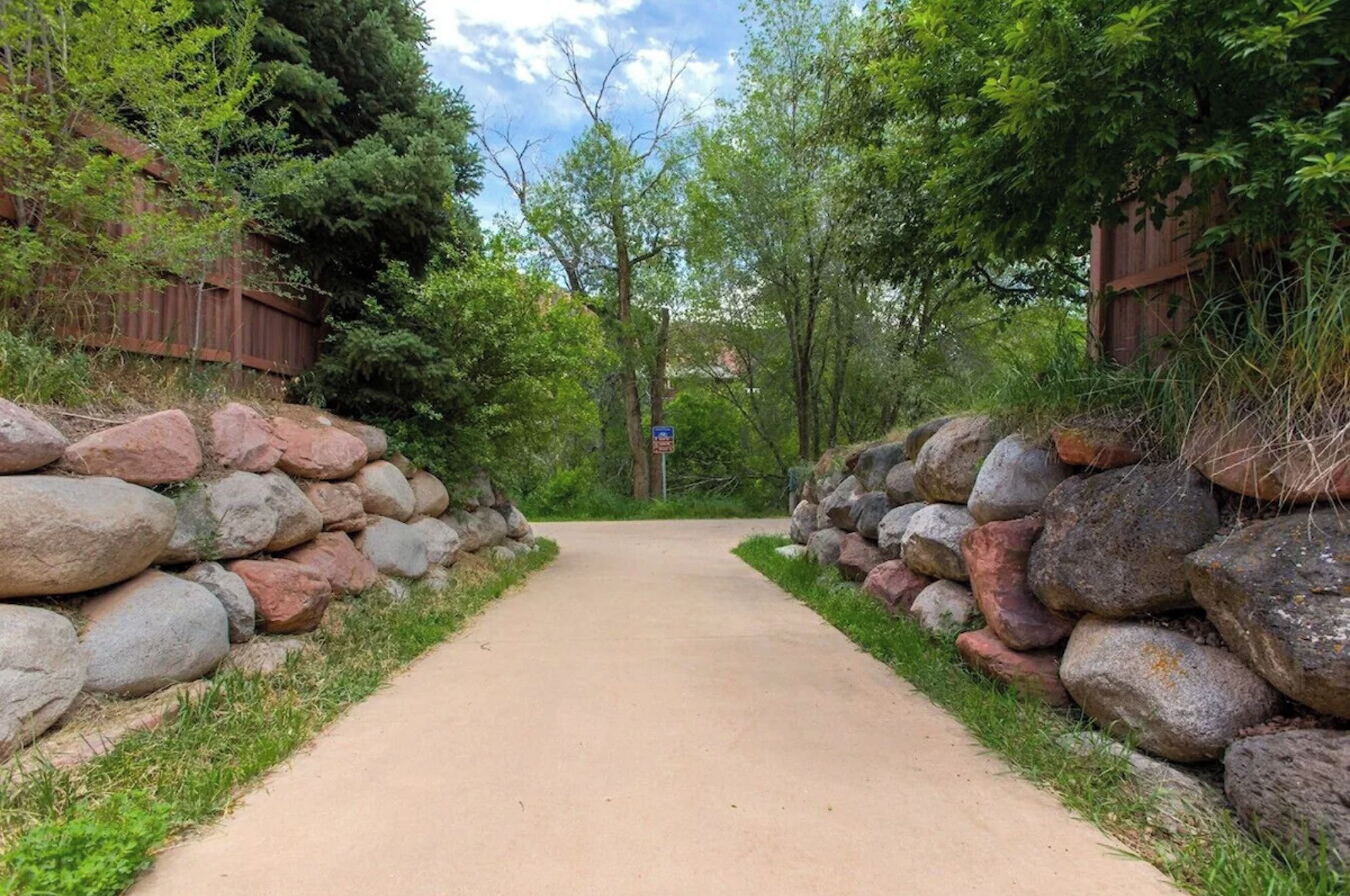 A paved pathway flanked by stone walls and dense foliage, leading into a wooded area.