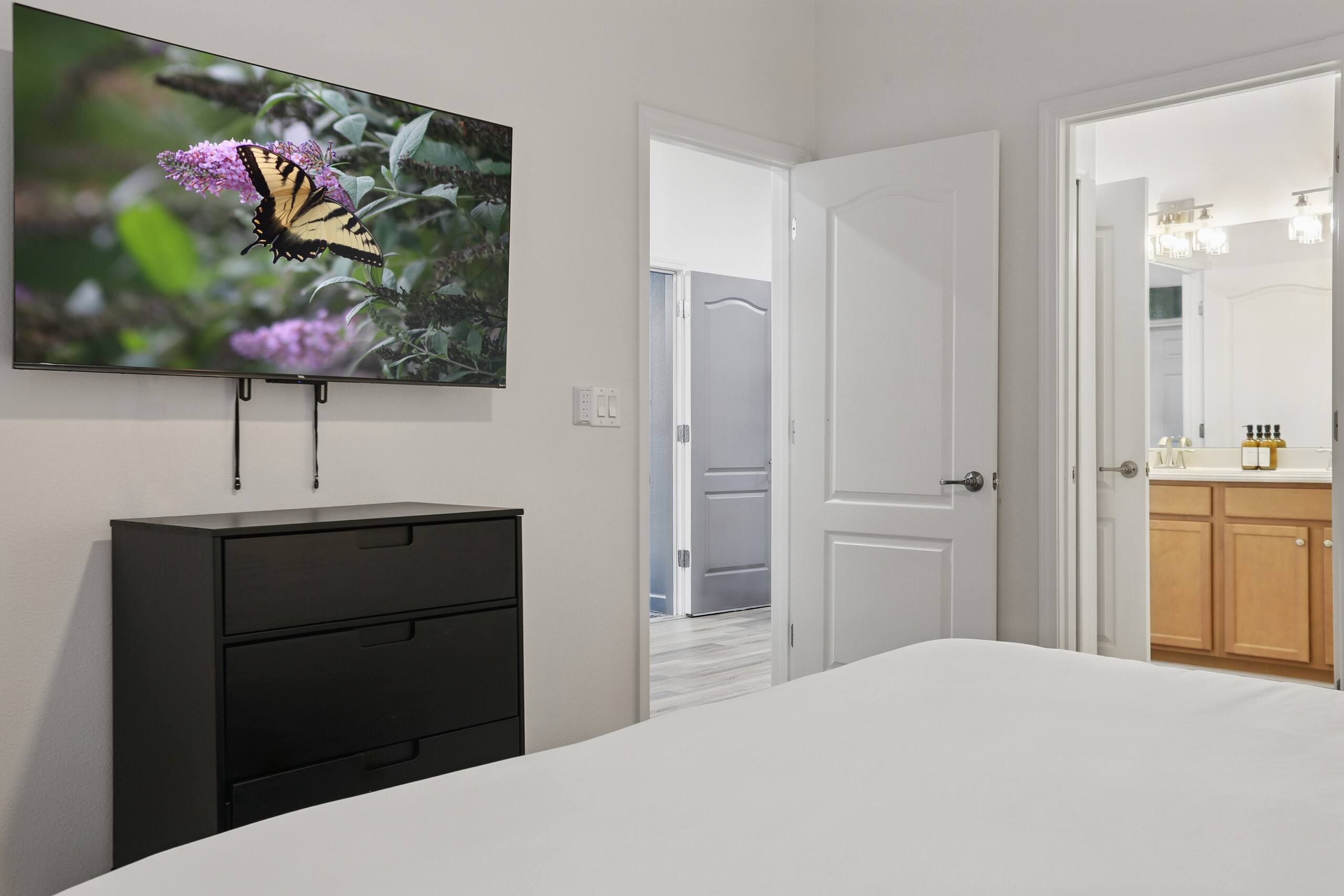 Une chambre à coucher comprenant un lit, une commode, un téléviseur fixé au mur et une porte donnant sur une autre pièce.