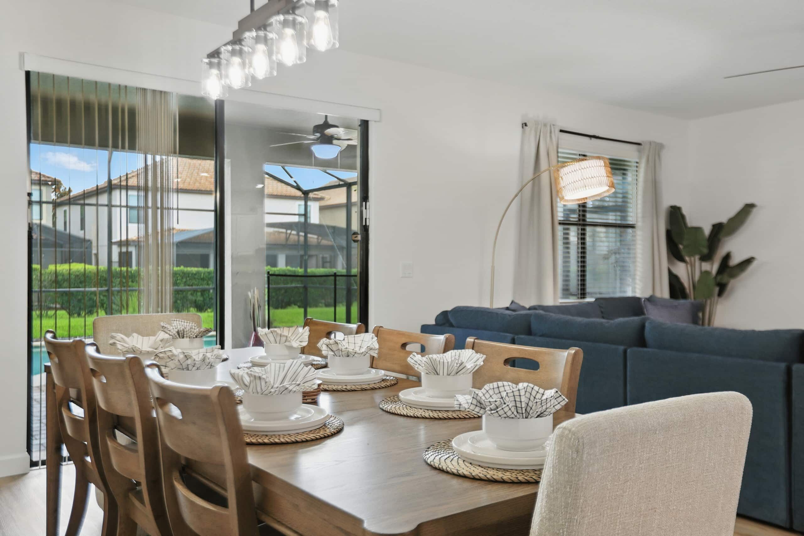 A dining area with a wooden table set for four, surrounded by chairs, with a view of a residential area through large windows.
