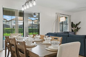 A dining area with a wooden table set for four, surrounded by chairs, with a view of a residential area through large windows. - Disney Vacation Pool Home | Resort | Game Room (Davenport)