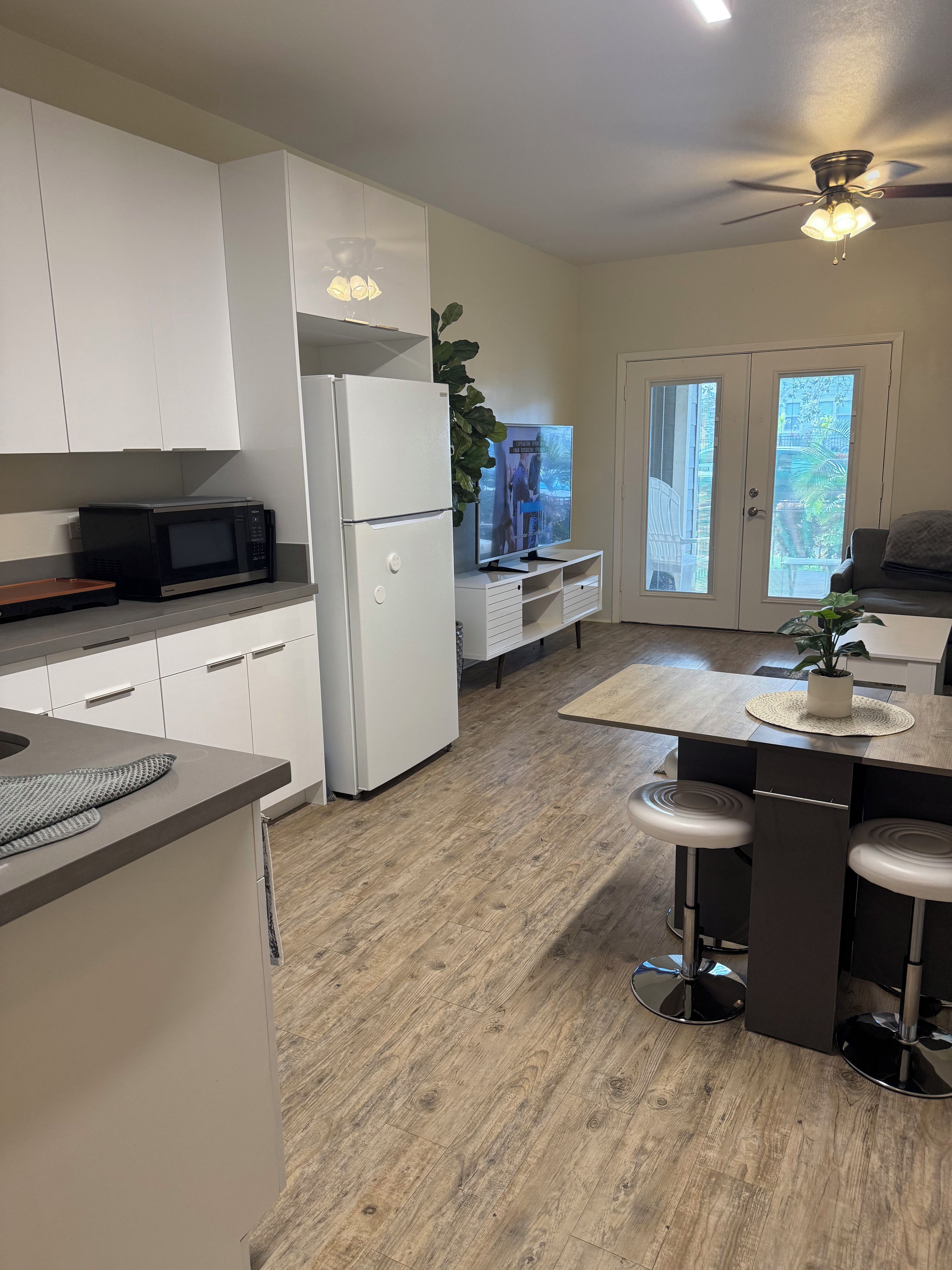 A modern kitchen with white cabinetry, a microwave, a refrigerator, a dining area with stools, and a ceiling fan.