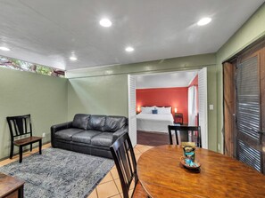 A room with a leather sofa, a dining table with chairs, a bed with red walls, and a window with blinds. - Authentic Eco-estate in Kona sleeps 14 -Living in Harmony with nature by the sea (Kailua-Kona)