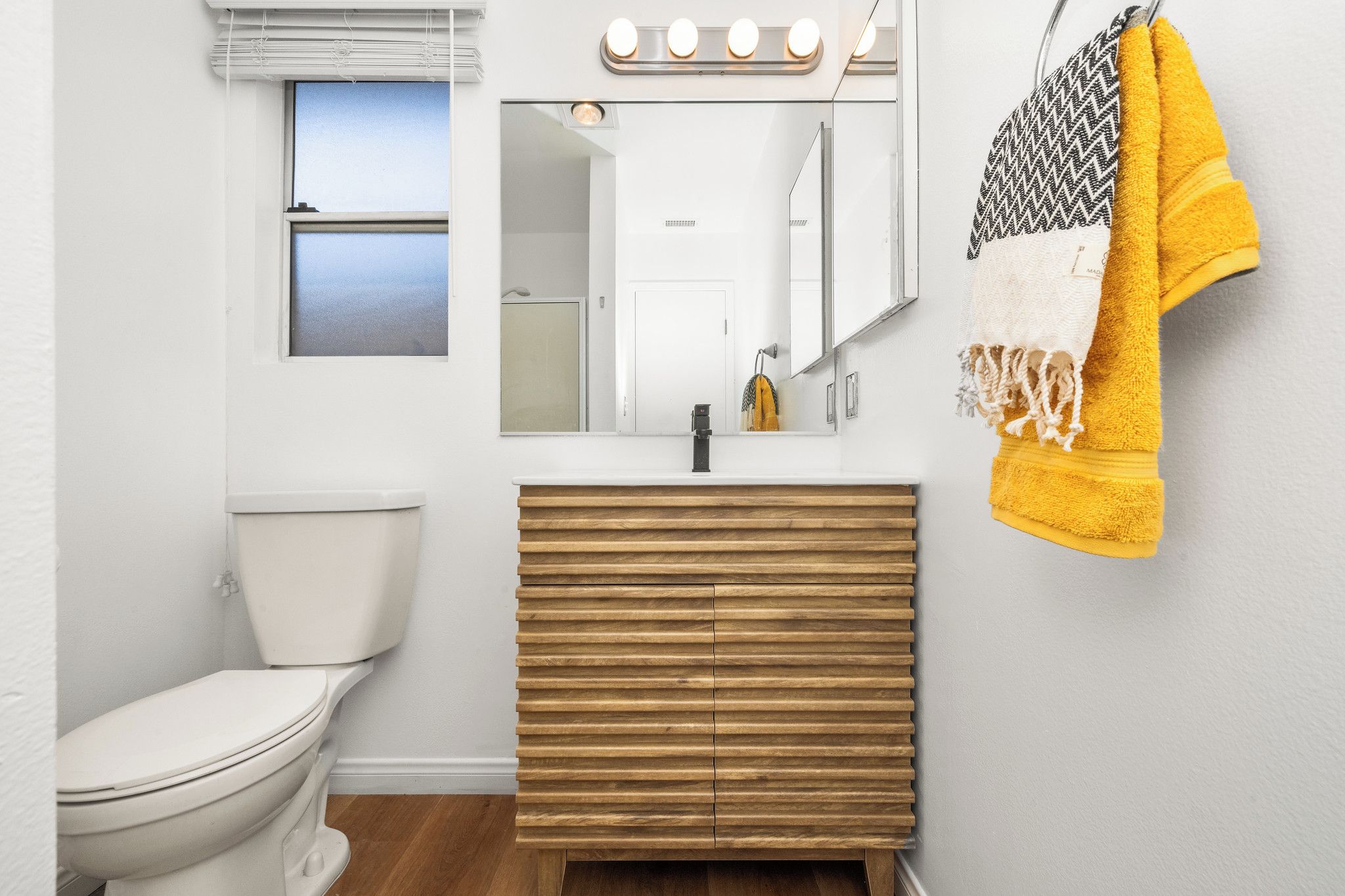 A bathroom with a toilet, a wooden vanity, a mirror with lights, and a towel rack.