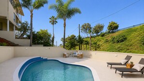 Ein Pool mit gewölbter Kante, umgeben von Liegestühlen und einem kleinen Tisch, mit Palmen und einer Hügelkette im Hintergrund.