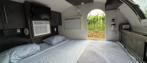 Dormitorio compacto de una caravana, con cama, ventilador de techo, ventana y vistas al exterior.
