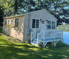 A small wooden cabin with a porch, located by a lake.