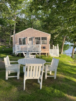 A wooden cabin with a deck, surrounded by trees and a grassy area with a table and chairs.