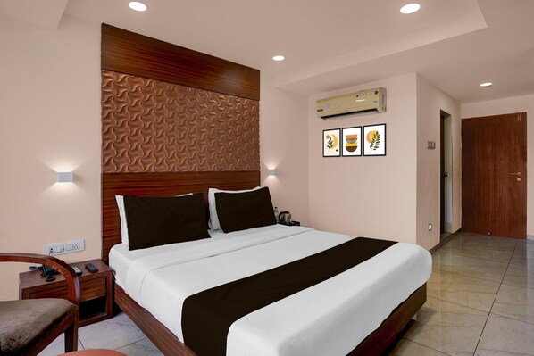 A hotel room with a large bed, a wooden headboard, a bedside table with a phone, a chair, and an air conditioning unit on the wall.