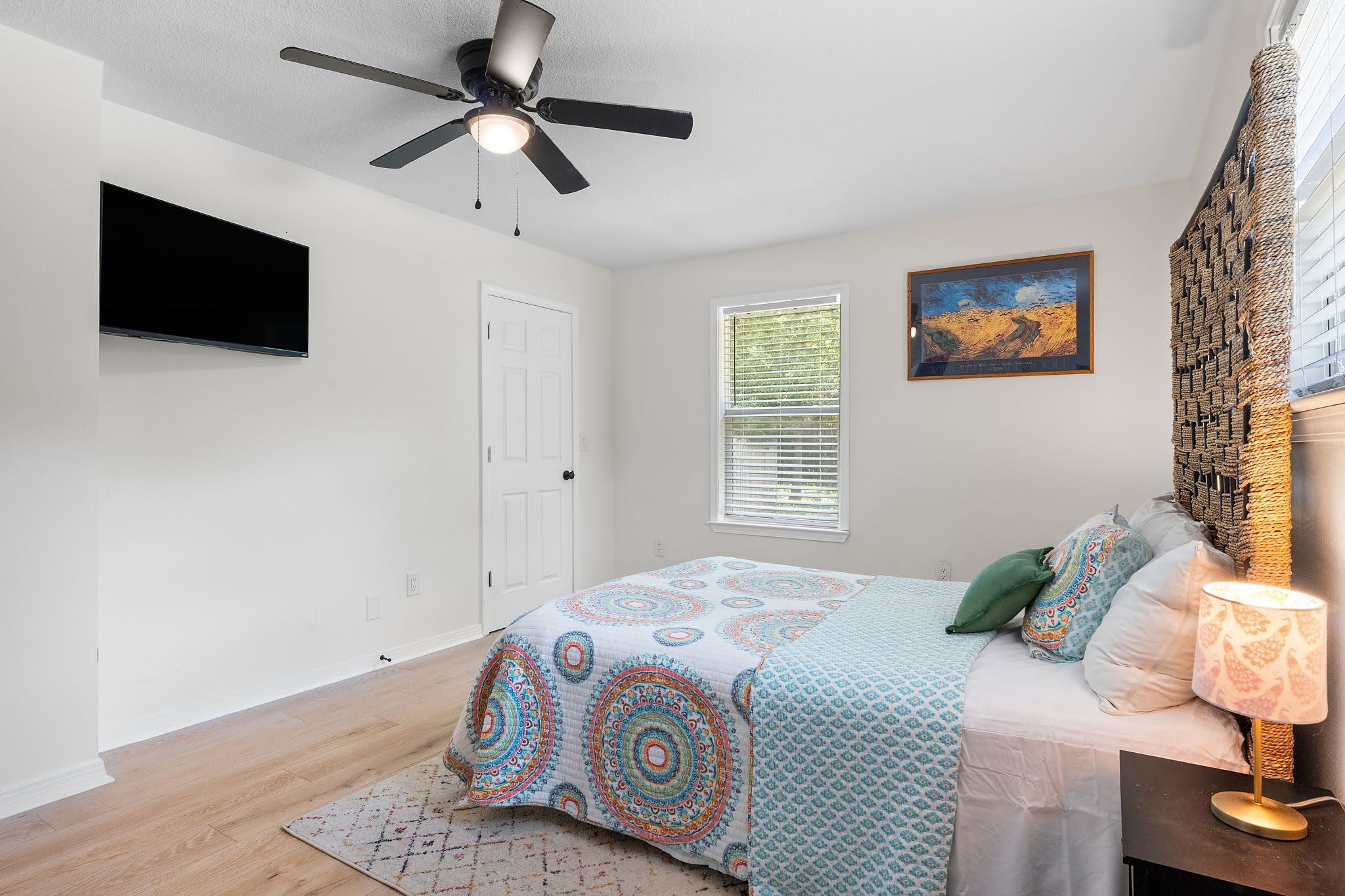 Une chambre à coucher avec un lit, un ventilateur de plafond, une fenêtre avec des stores, un tableau et une lampe.